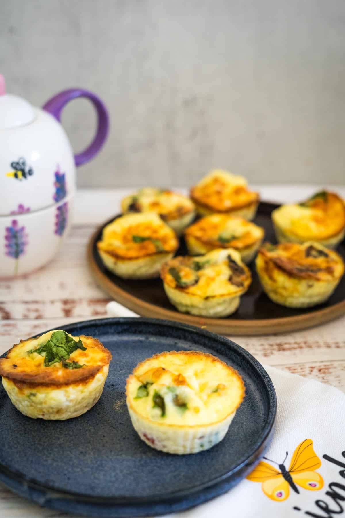 Two small quiches on a dark blue plate with more quiches on a dark tray in the background. A partially visible ornate teapot sits to the left. A cloth with a butterfly design is in the foreground, reminiscent of cottage cheese egg muffins from a cozy kitchen.