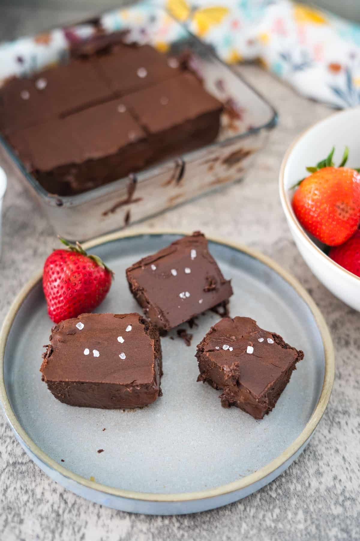 Plate with three chocolate fudge squares, sprinkled with sea salt, next to a bowl of strawberries. A larger glass dish with additional cottage cheese brownies is in the background.