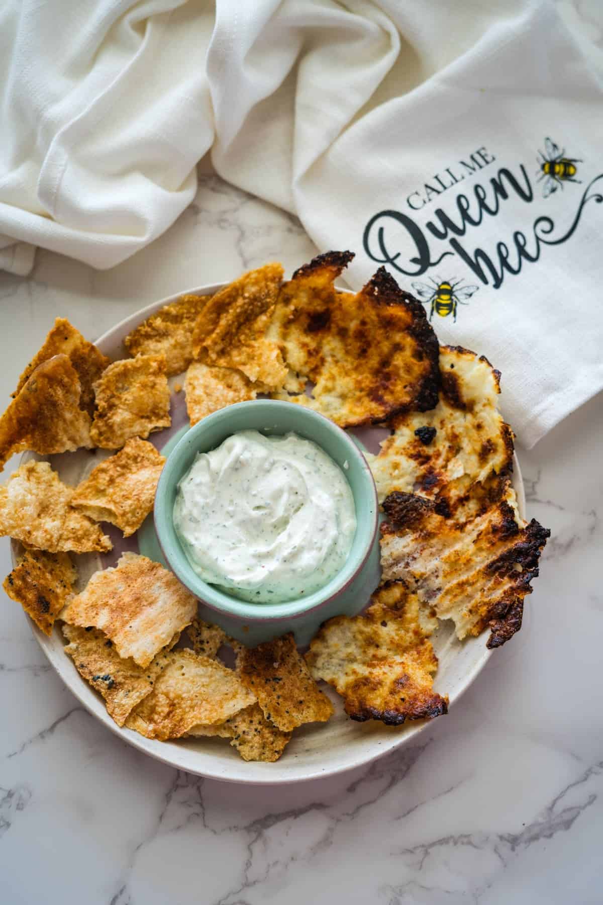 A plate of crispy bread slices surrounds a small bowl of white creamy dip, reminiscent of a luscious blend like cottage cheese. In the background, a napkin with the text "Call Me Queen Bee" and bee illustrations adds a whimsical touch.