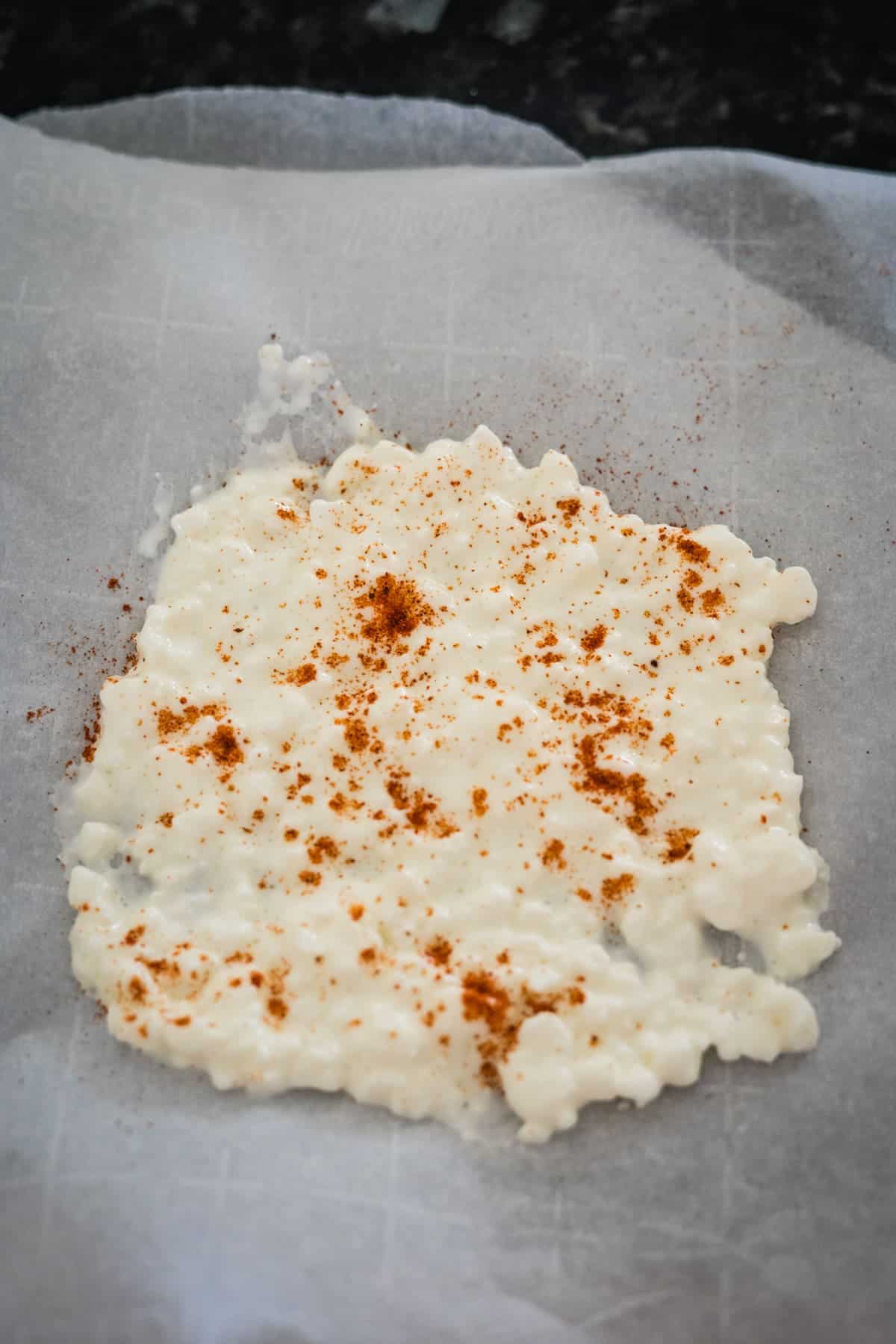 Parchment paper with white melted cheese sprinkled with a small amount of paprika, on a dark countertop.