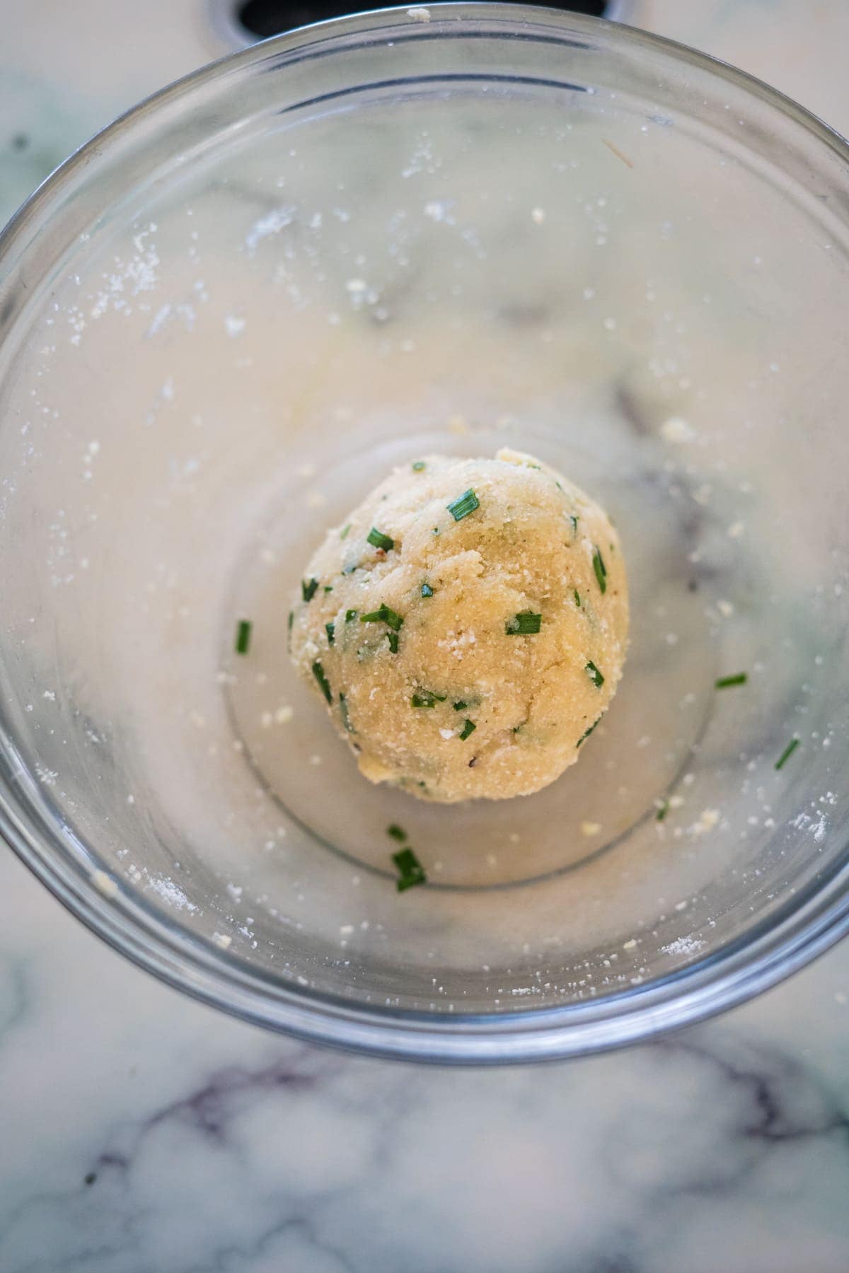 A ball of dough with chopped herbs is in a clear glass bowl on a marble surface.