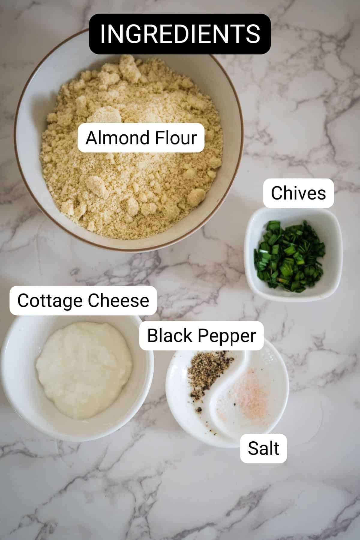 A bowl of almond flour, chopped chives, cottage cheese, black pepper, and salt in small dishes, labeled as ingredients on a marble countertop.