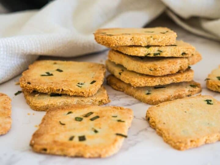 A stack of square cottage cheese crackers with green herbs rests on a white surface next to a white cloth. Some crackers are spread around in the foreground.