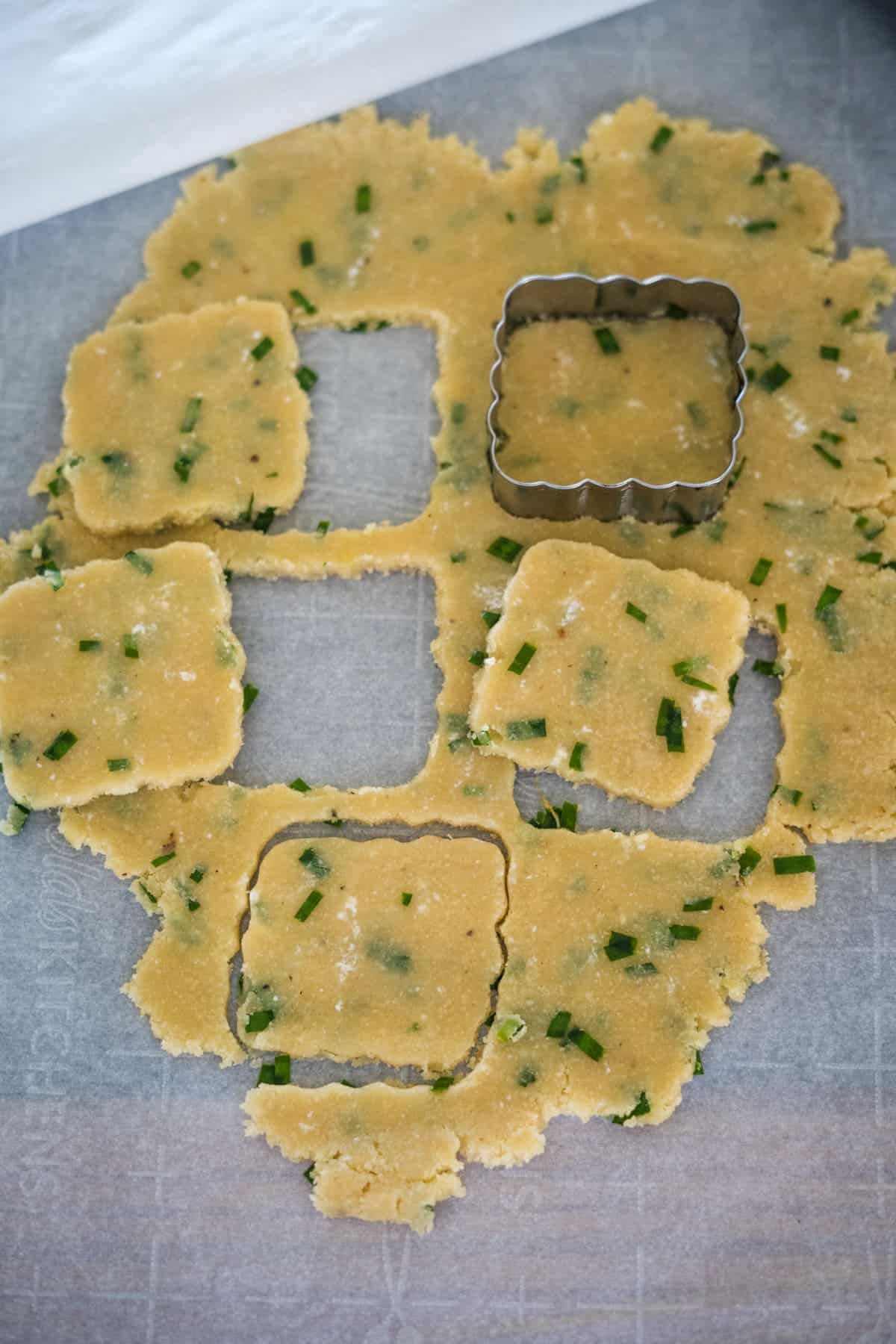 A sheet of rolled-out dough with green herbs, partially cut into square shapes using a square cookie cutter, laid on parchment paper. Some cut-out squares are arranged around the dough.