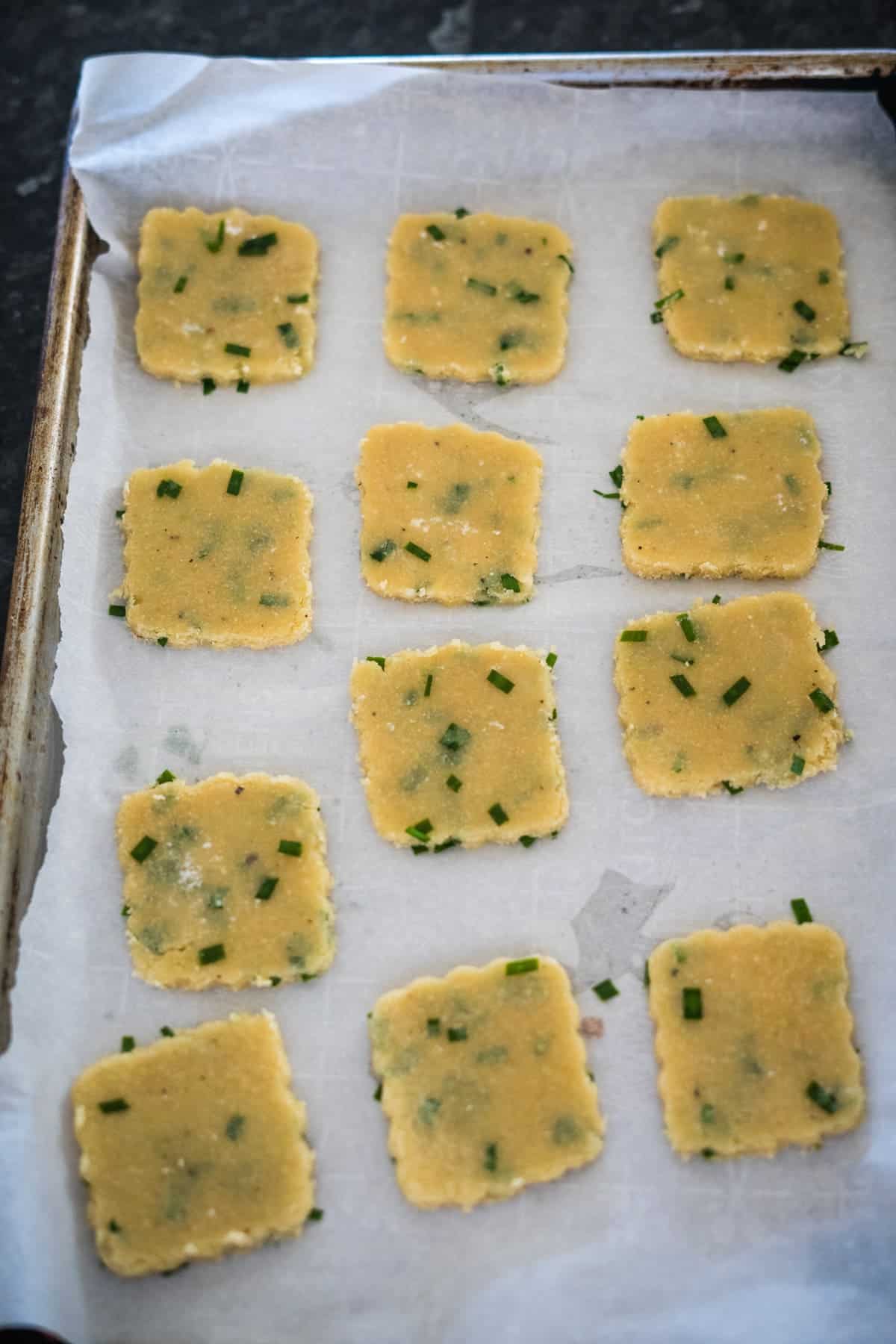 A baking tray with 12 uncooked square crackers dotted with green herbs, arranged in three rows, placed on parchment paper.