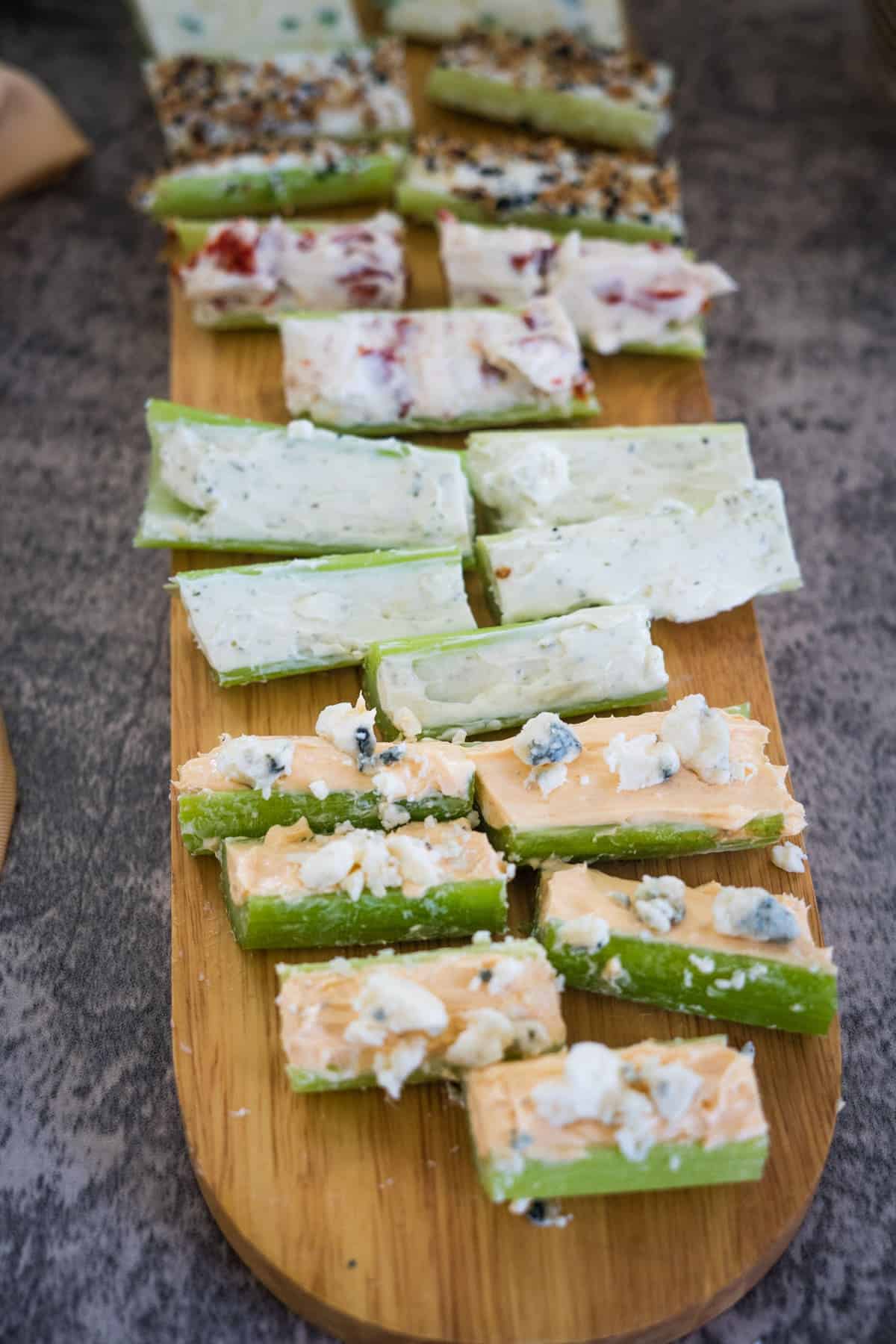 A wooden board displaying several rows of celery sticks filled with various toppings, including cream cheese stuffed celery and other spreads, is set against a textured stone surface.