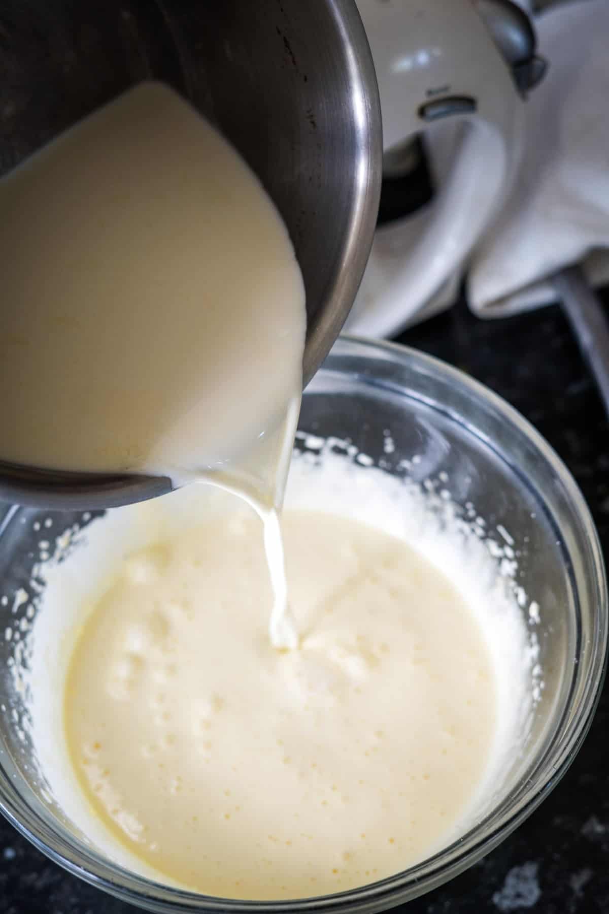 A metal pot pours liquid into a glass bowl containing a creamy mixture, creating the perfect base for keto vanilla ice cream.