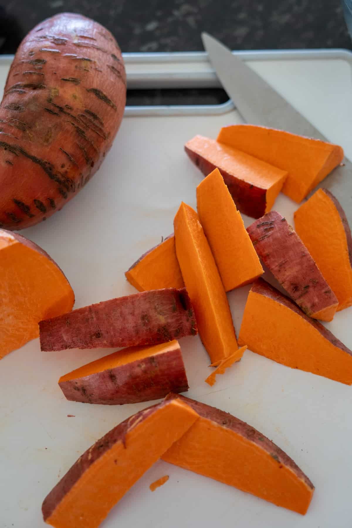 A partially peeled sweet potato on a white cutting board, next to a knife and several orange sweet potato slices.