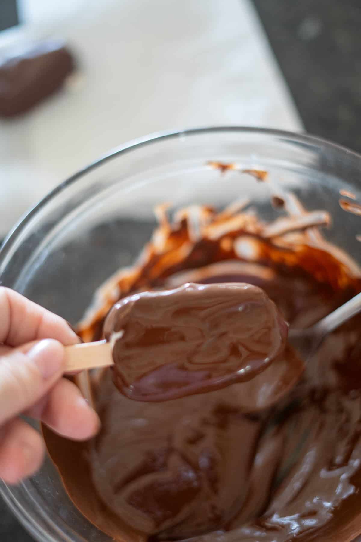 A hand holding a chocolate-covered keto ice cream bar over a bowl filled with melted chocolate.