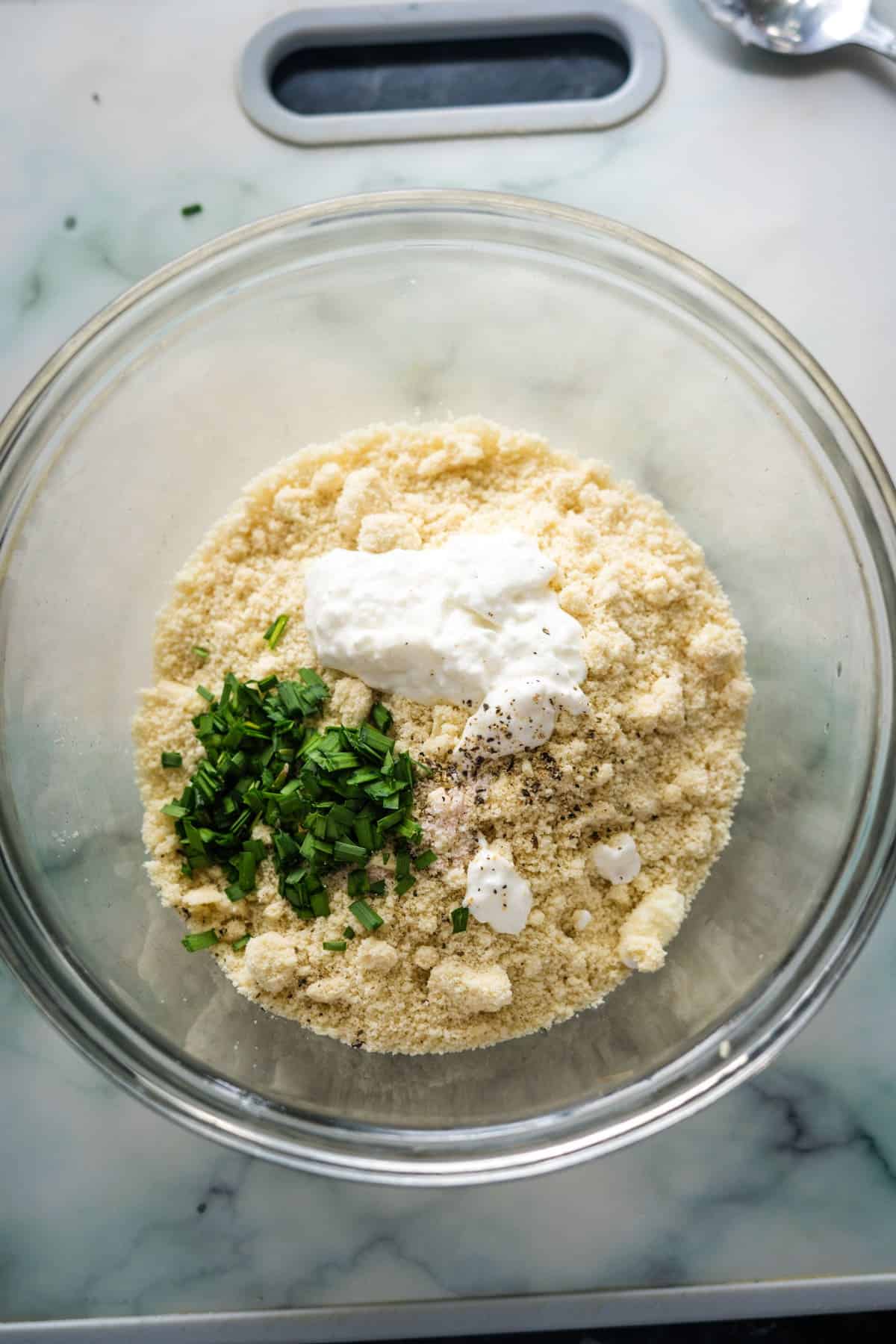 A glass bowl contains a mixture of almond flour, chopped green herbs, black pepper, and dollops of white cream or yogurt, placed on a marble countertop near a silver spoon.