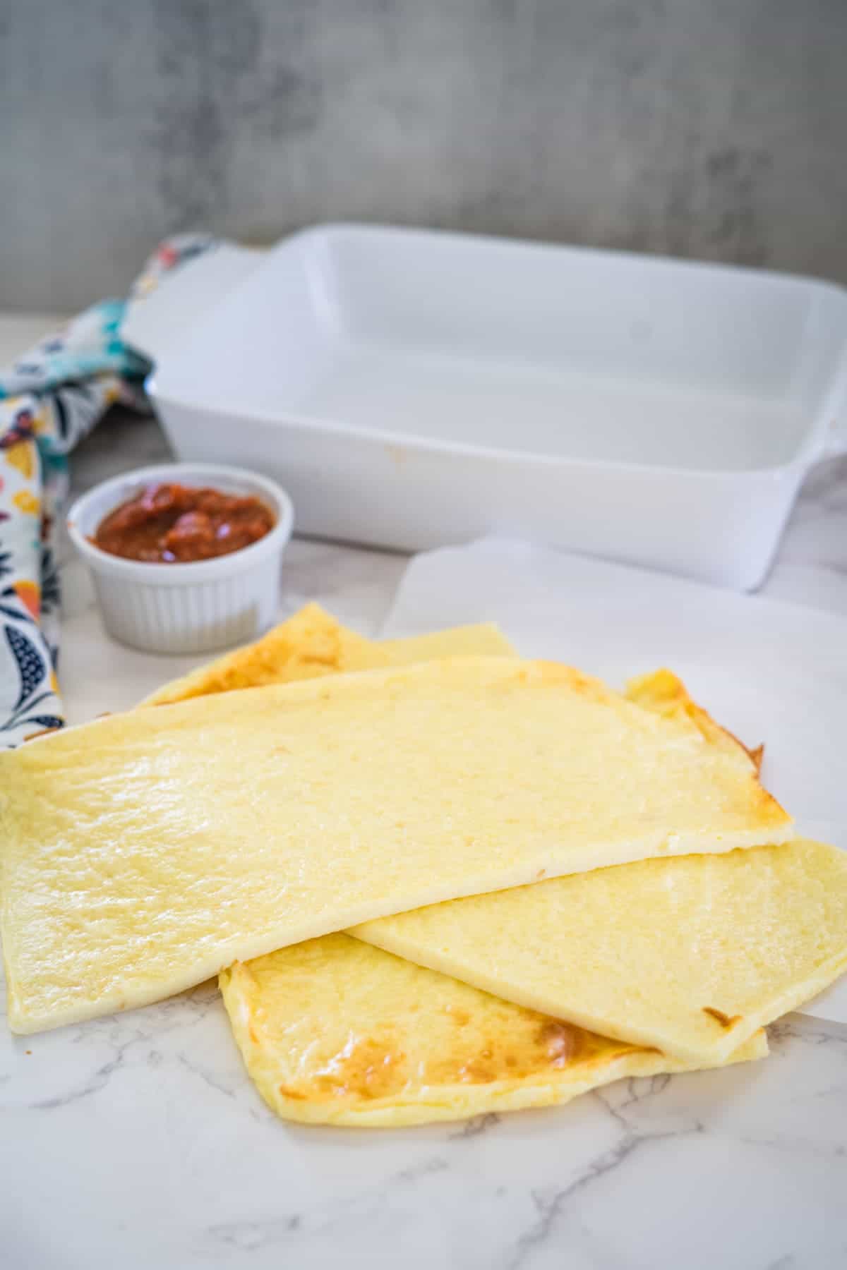 Several sheets of homemade keto lasagna noodles are laid out on a marble surface next to a small bowl of sauce and an empty white baking dish in the background.