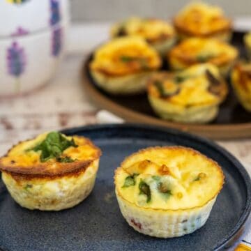 Close-up of two mini quiches garnished with herbs on a blue plate, with more mini quiches on a tray in the background.