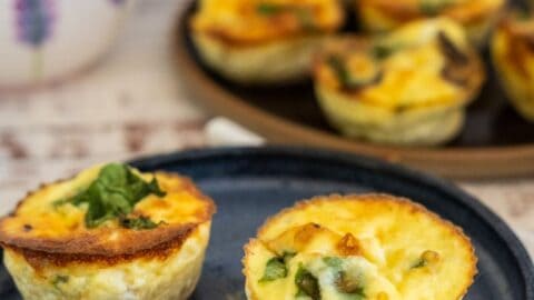 Close-up of two mini quiches garnished with herbs on a blue plate, with more mini quiches on a tray in the background.