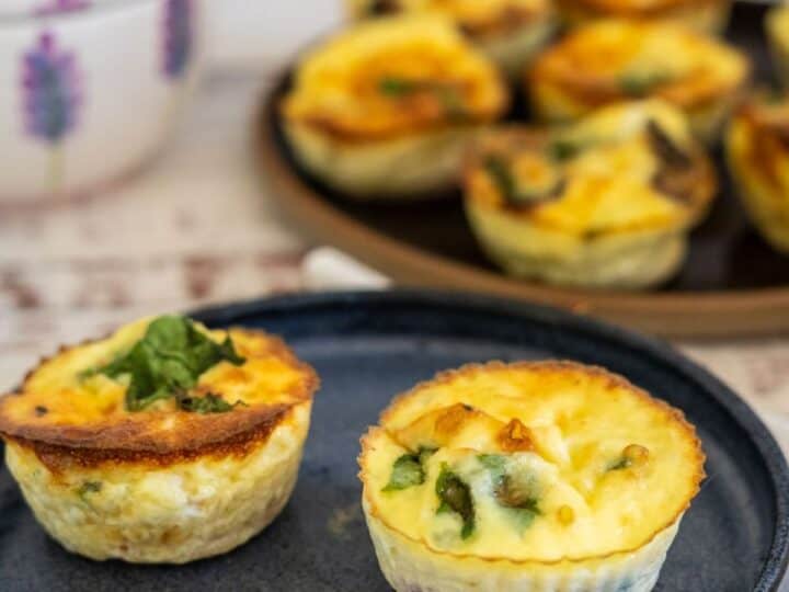 Close-up of two mini quiches garnished with herbs on a blue plate, with more mini quiches on a tray in the background.