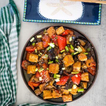 A plate of stir-fried tofu with red bell peppers, dark leafy greens, and green onions, garnished with sesame seeds, next to a green checked towel.