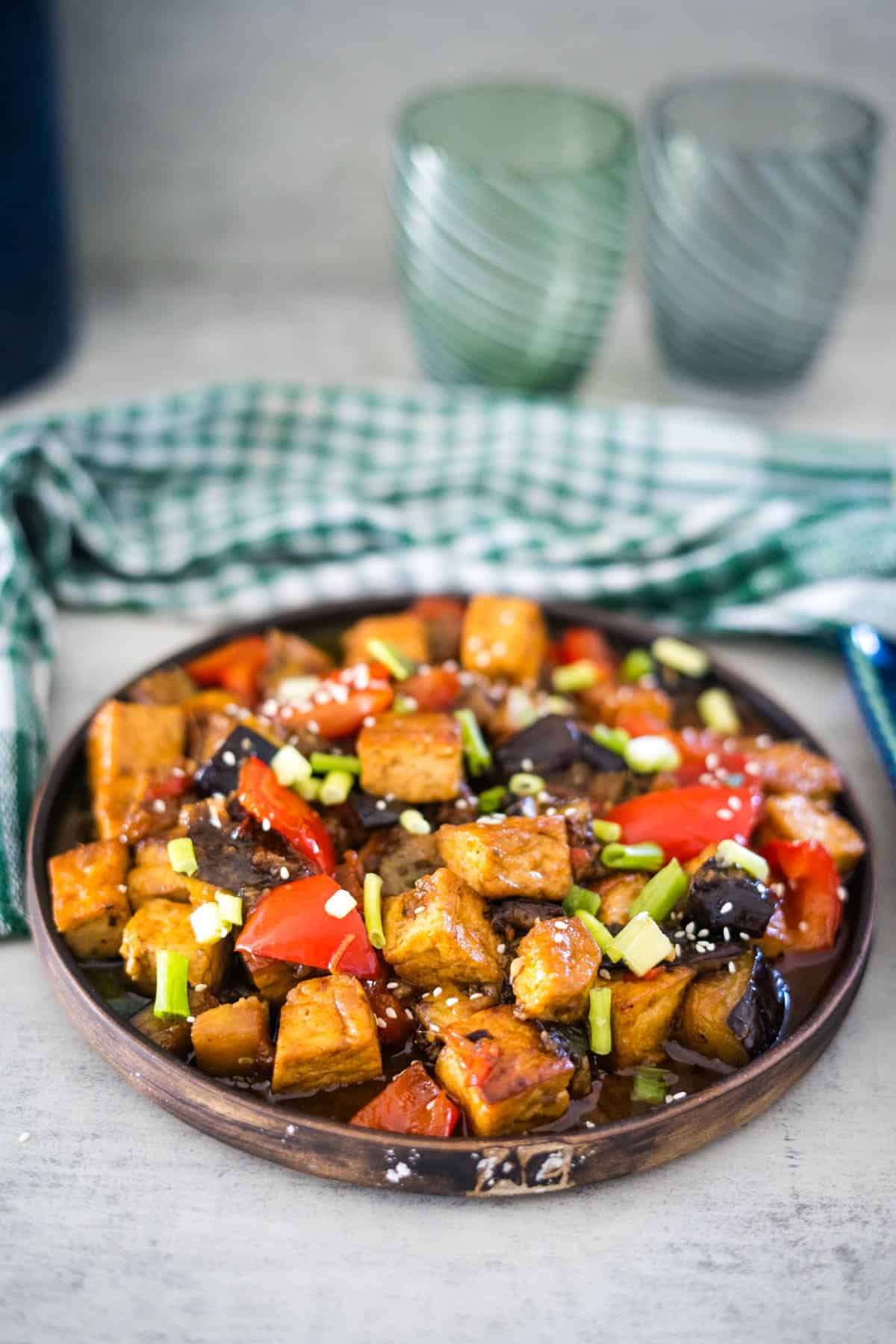 A wooden plate filled with a stir-fried dish featuring tofu cubes, red bell peppers, eggplant, green onions, and sesame seeds. Two green glass cups and a green checkered cloth are in the background.