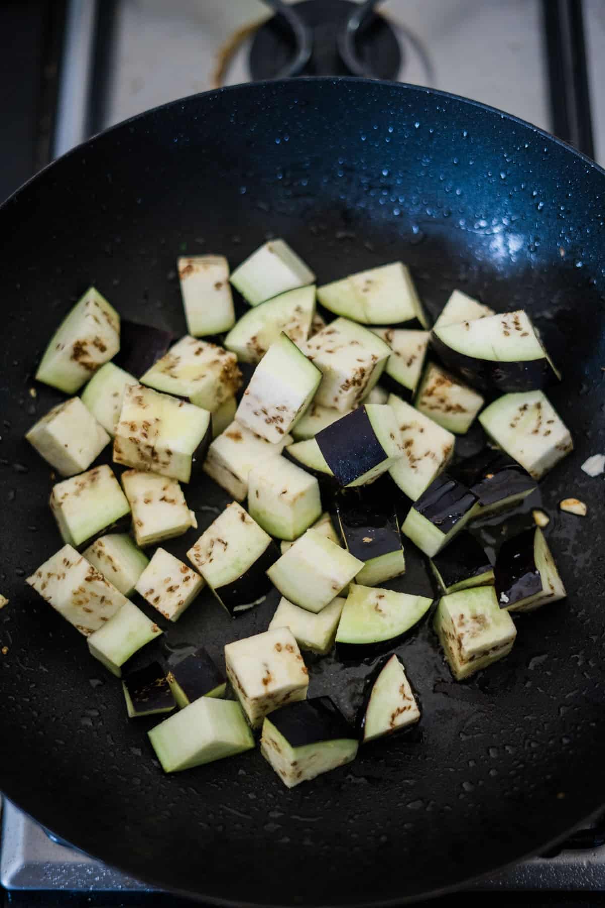 Cubed eggplant pieces are being cooked in a black frying pan on a stove, showcasing a delicious eggplant tofu recipe.