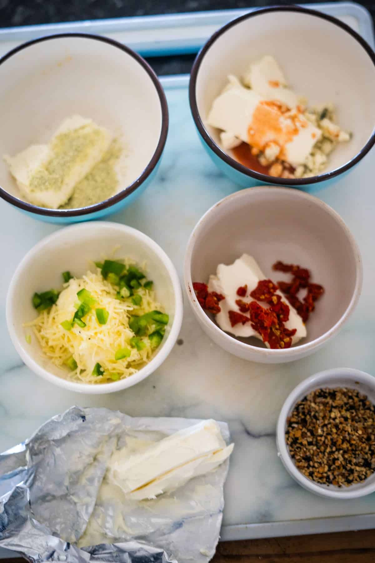 An assortment of ingredients in bowls on a countertop: a bowl with cheese and green peppers, another with sun-dried tomatoes and cream cheese, a third with seasoning and cream cheese, a bowl of seeds, and a plate of cream cheese stuffed celery.