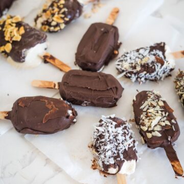 Assorted chocolate-covered ice cream bars on sticks, topped with various toppings such as nuts, coconut flakes, and chocolate drizzle, placed on a white paper-lined surface.