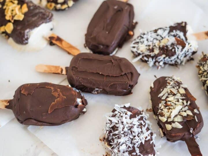 Assorted chocolate-covered ice cream bars on sticks, topped with various toppings such as nuts, coconut flakes, and chocolate drizzle, placed on a white paper-lined surface.