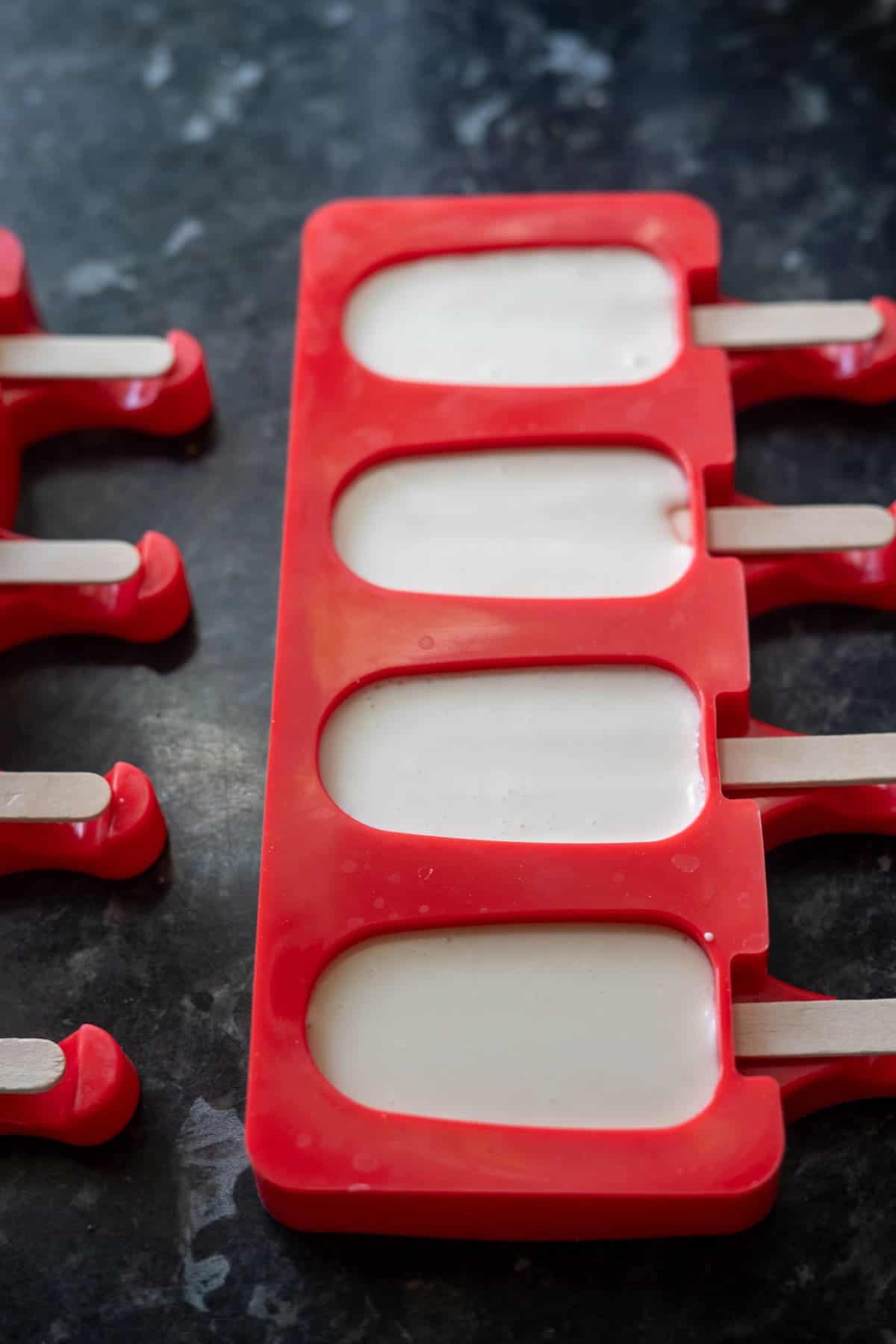 Red popsicle mold filled with white liquid, positioned on a dark surface. Wooden sticks are inserted into the mold, ready to transform this concoction into delicious keto ice cream bars.