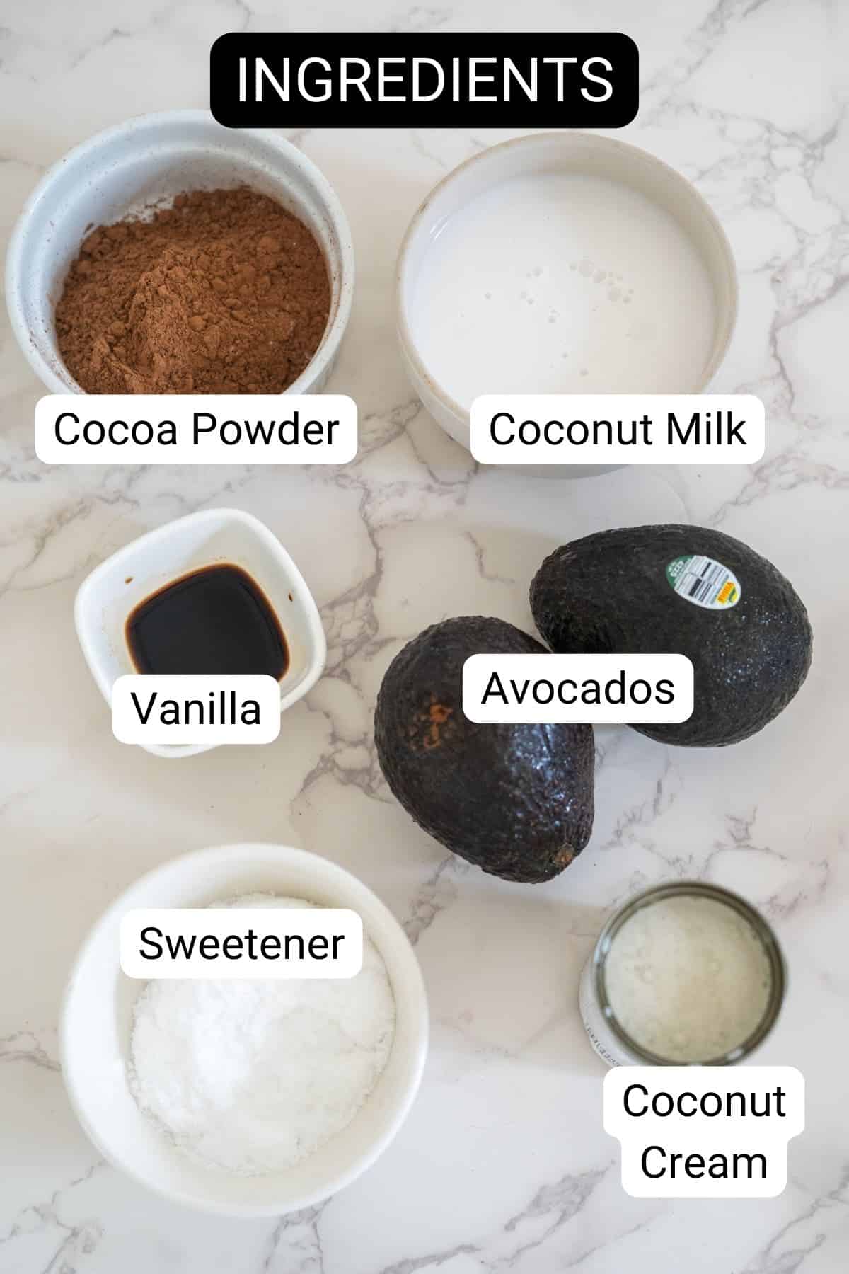 Ingredients for a recipe, labeled and arranged on a marble countertop: cocoa powder, coconut milk, vanilla, avocados, sweetener, and coconut cream.