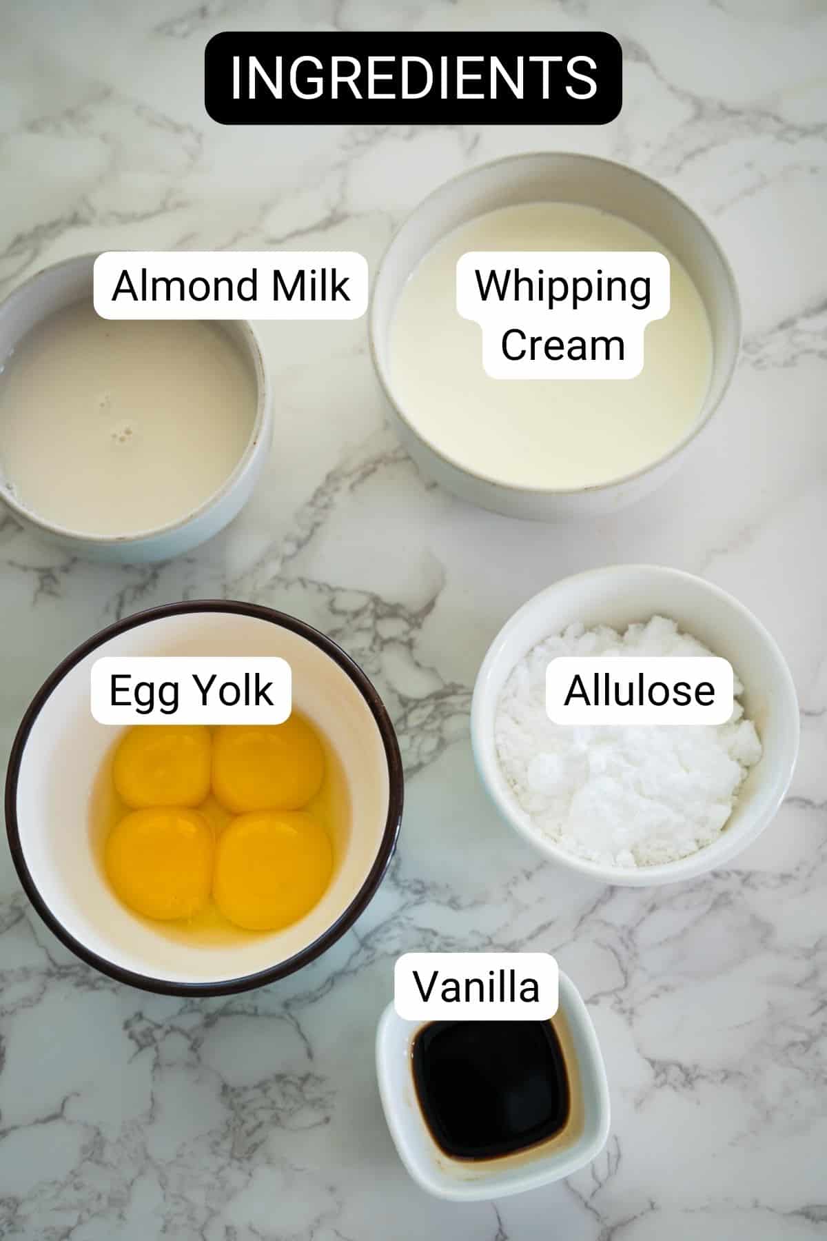 Ingredients for keto vanilla ice cream on a marble surface include bowls of almond milk, whipping cream, egg yolks, allulose, and vanilla extract.