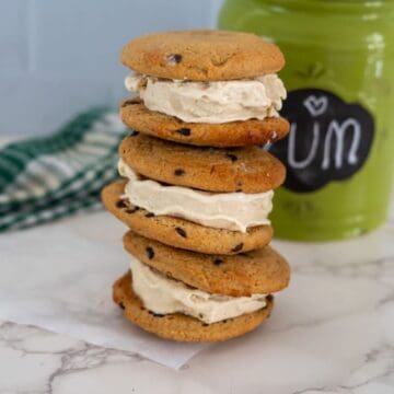 A stack of three chocolate chip cookie ice cream sandwiches on a marble surface with a green jar labeled "Yum" in the background.