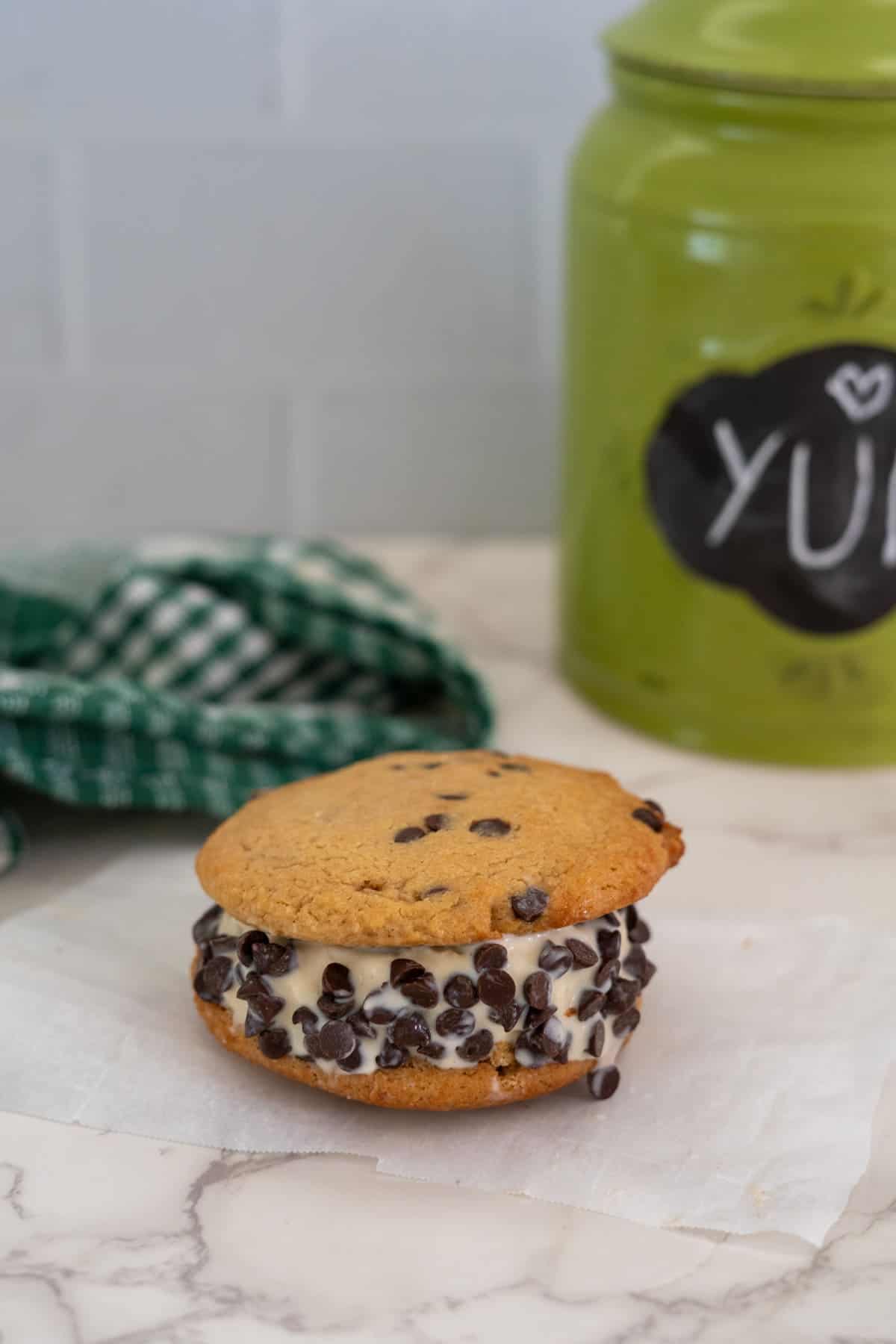 A chocolate chip cookie ice cream sandwich with chocolate chips, placed on parchment paper, with a green checkered cloth and a green jar labeled "YUM" in the background.