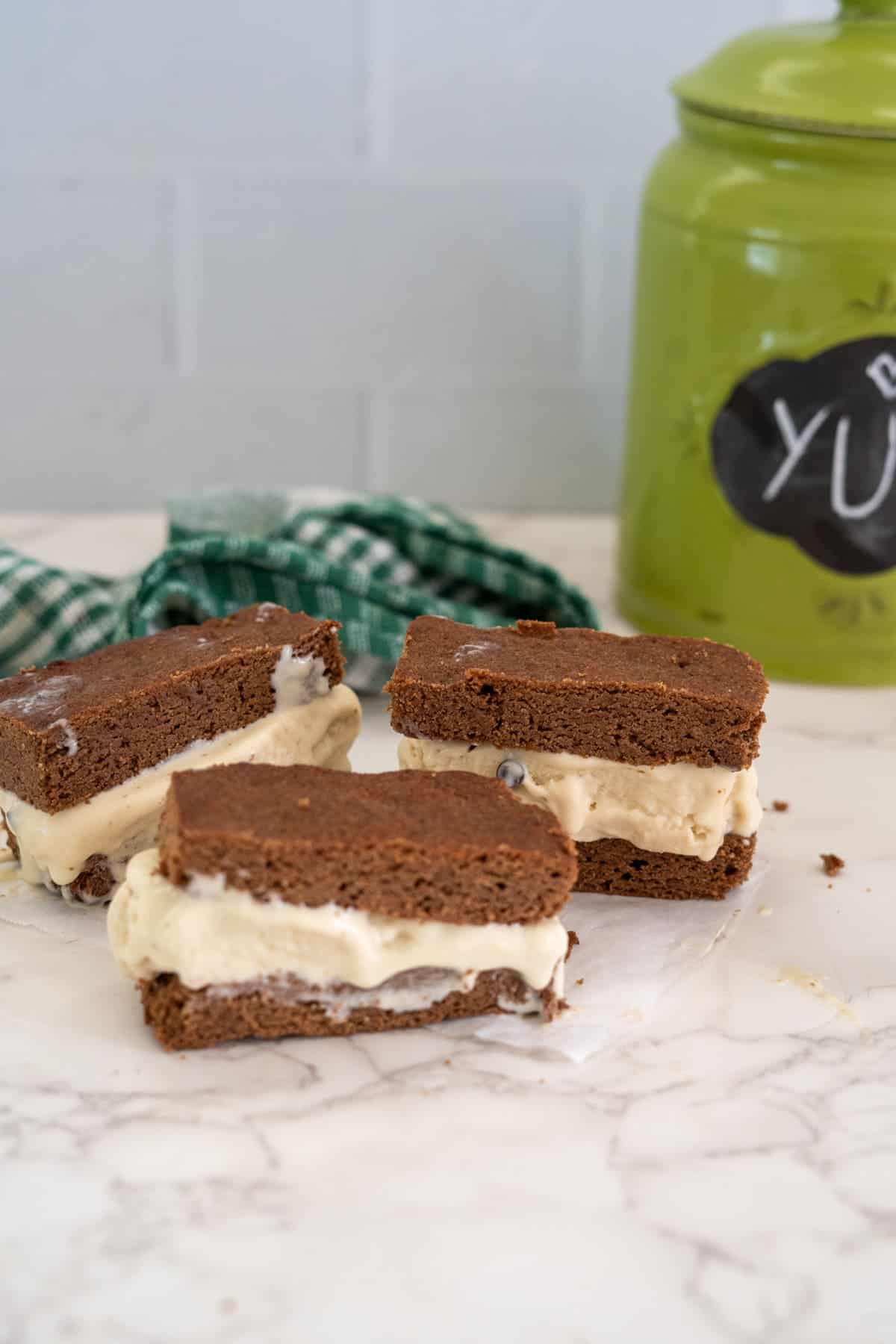 Three keto ice cream sandwiches made with chocolate chip cookies and vanilla ice cream are placed on a marble surface. In the background, there is a green container labeled "Yum" and a green checkered cloth.