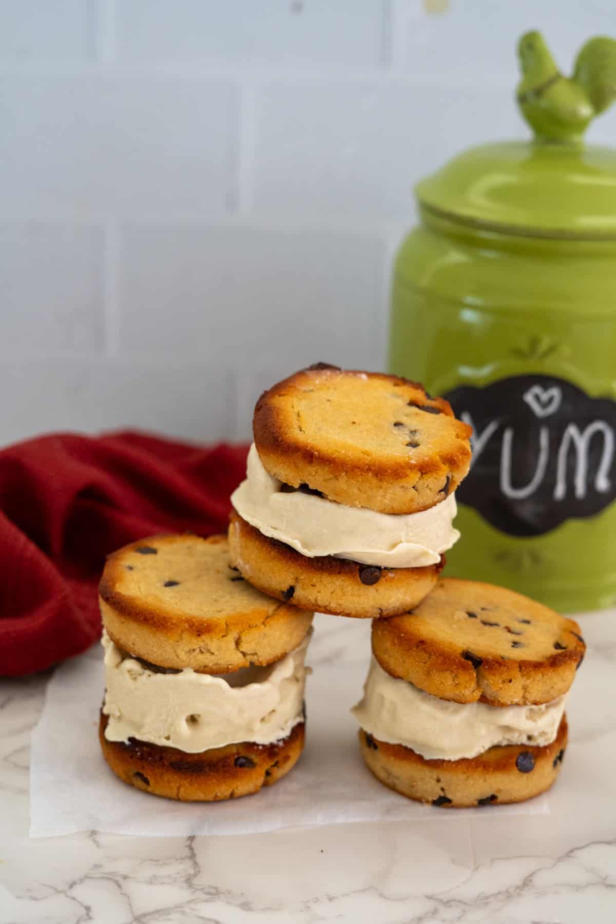 Three keto chocolate chip ice cream sandwiches stacked on a marble countertop with a green container and a red cloth in the background.