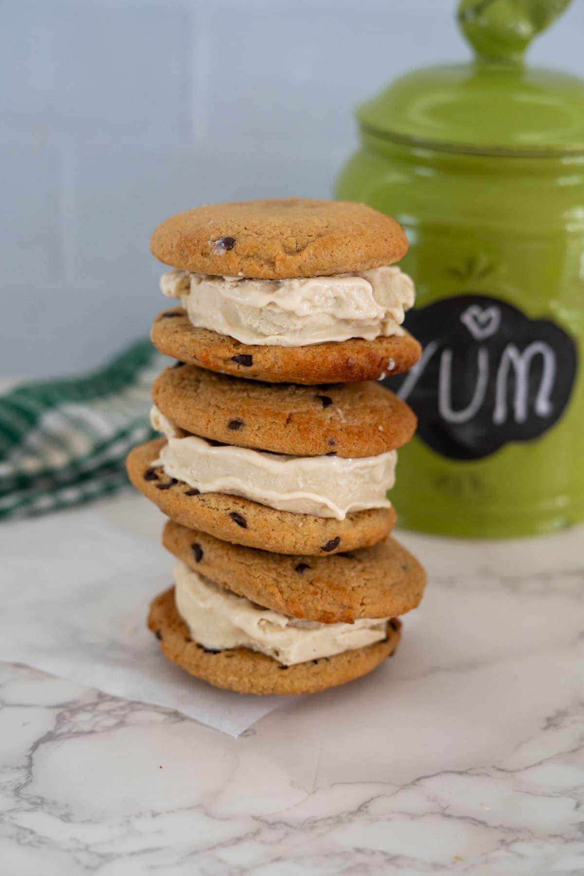 A stack of four ice cream sandwiches made with keto chocolate chip cookies is placed on a marble surface. A green container with "YUM" written in chalk is in the background.