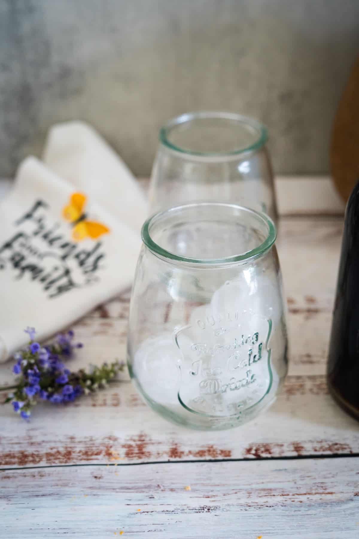 Two empty glasses on a wooden surface, a small bouquet of purple flowers, a white cloth with text, and part of a dark bottle in the background.