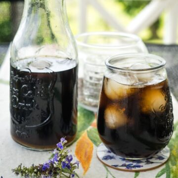 Glass bottle and a small glass filled with iced cola on a floral tablecloth; a purple flower is visible in the foreground.