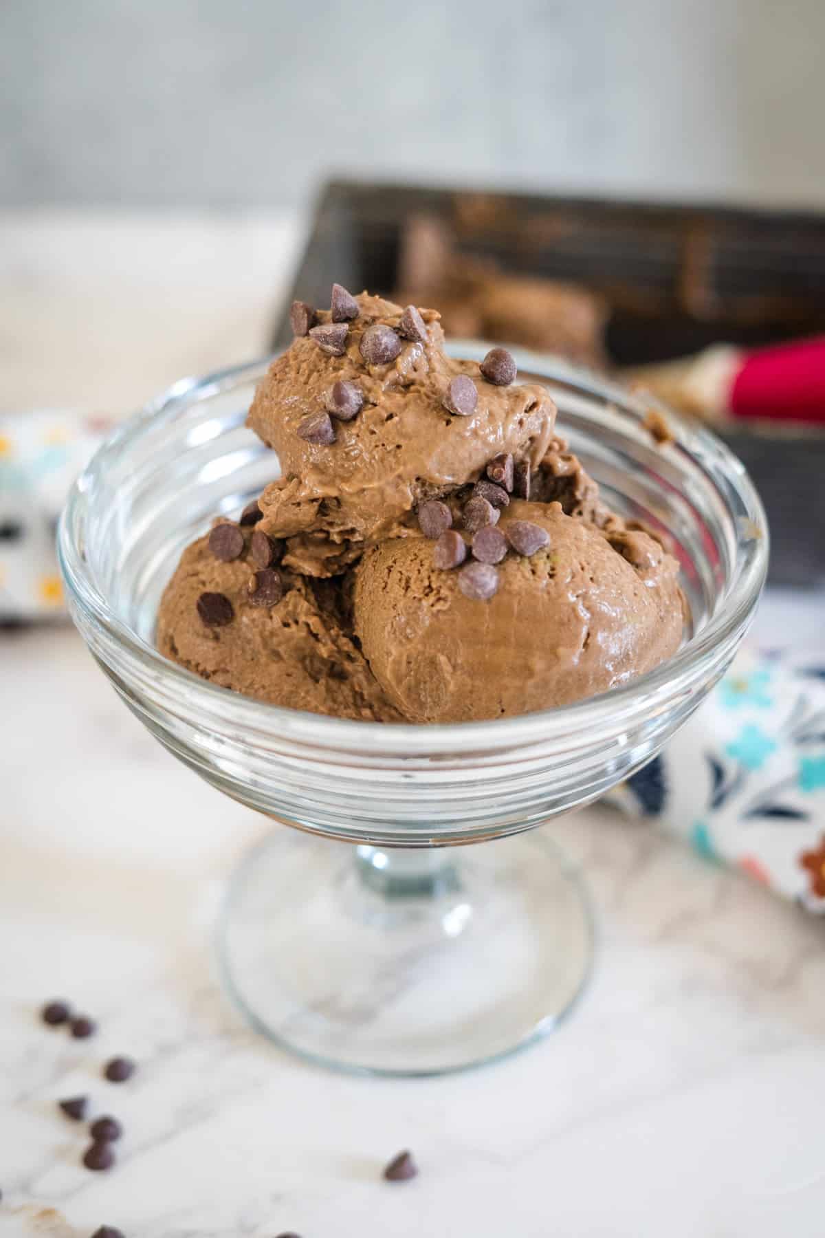 A glass bowl filled with three scoops of chocolate ice cream, garnished with chocolate chips, sits on a marble surface.