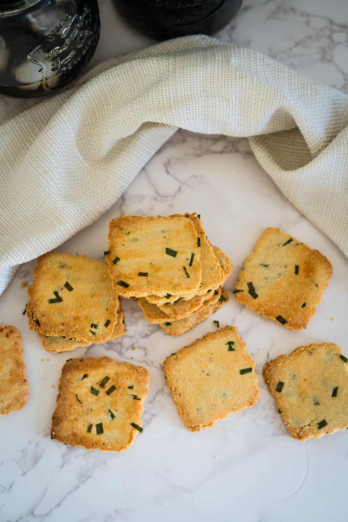 A stack of square, golden-brown cottage cheese crackers with green herbs sits on a marble surface next to a cloth and two jars.