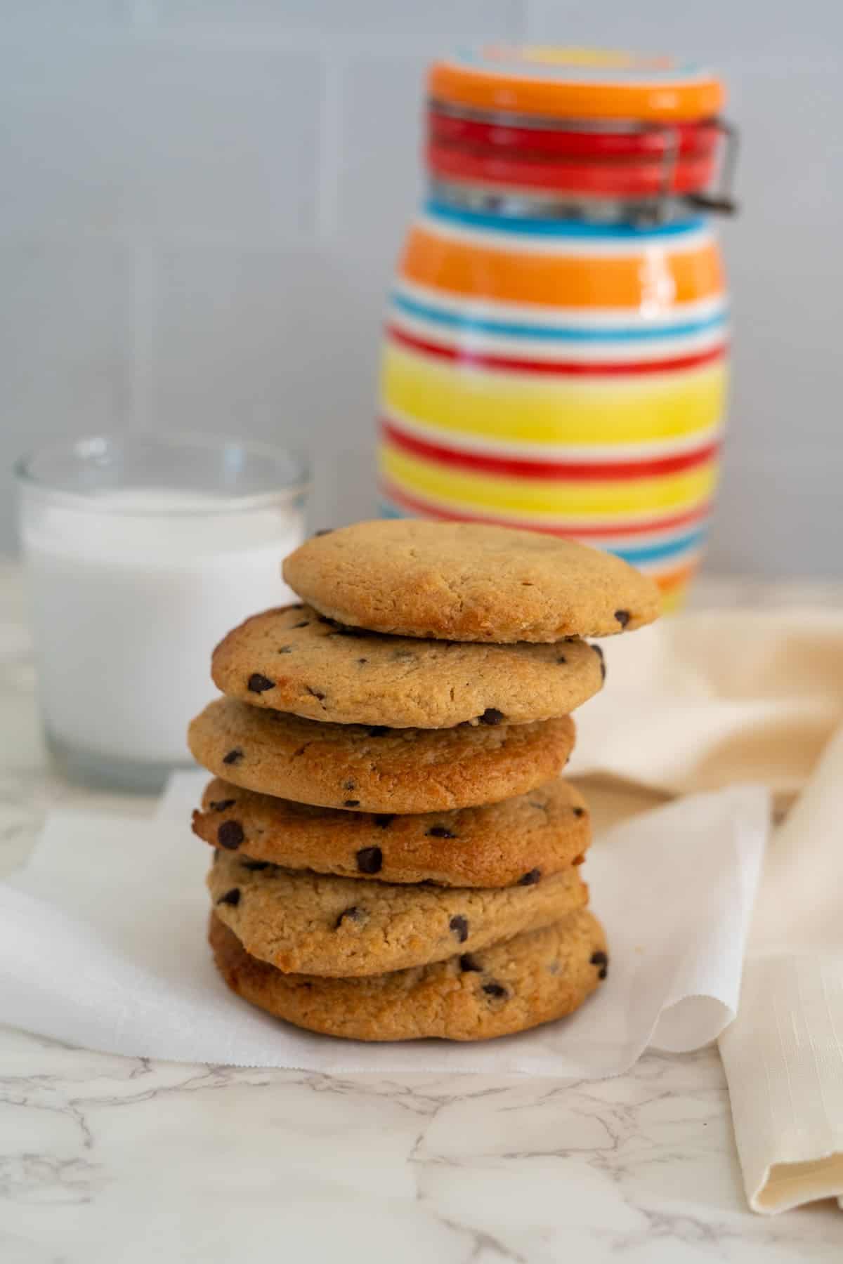 A stack of cookies with chocolate chips on a white napkin, a glass of milk, and a colorful striped jar in the background on a marble surface.