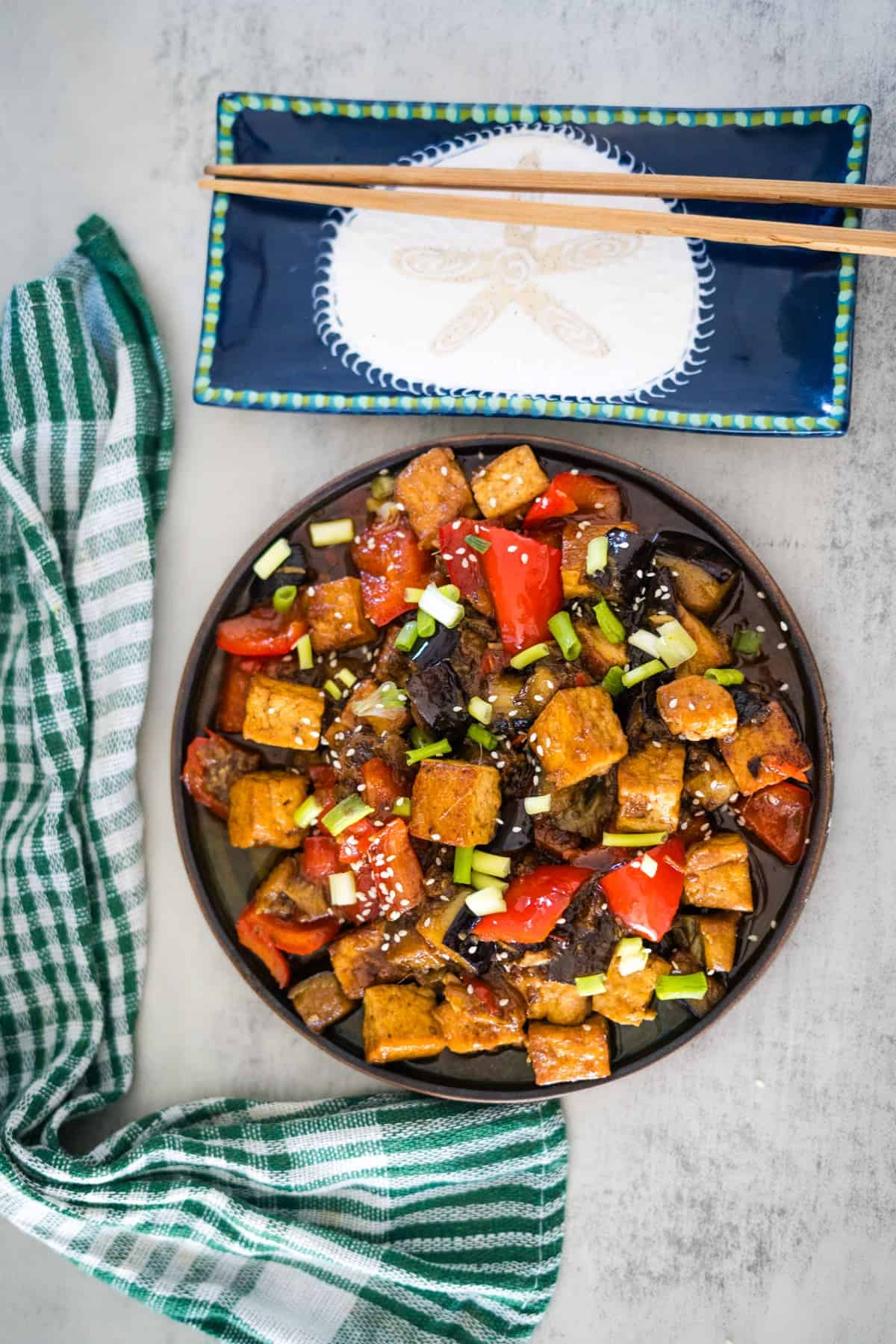 A bowl of stir-fried tofu with red bell peppers, onions, green onions, and tender eggplant, displayed on a grey surface with chopsticks on a ceramic dish and a green-checked cloth beside it.