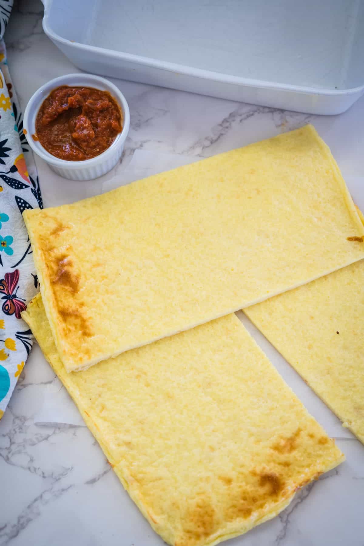 Two rectangular cooked pasta sheets placed on a marble countertop next to a small white ramekin filled with red sauce and a colorful cloth.