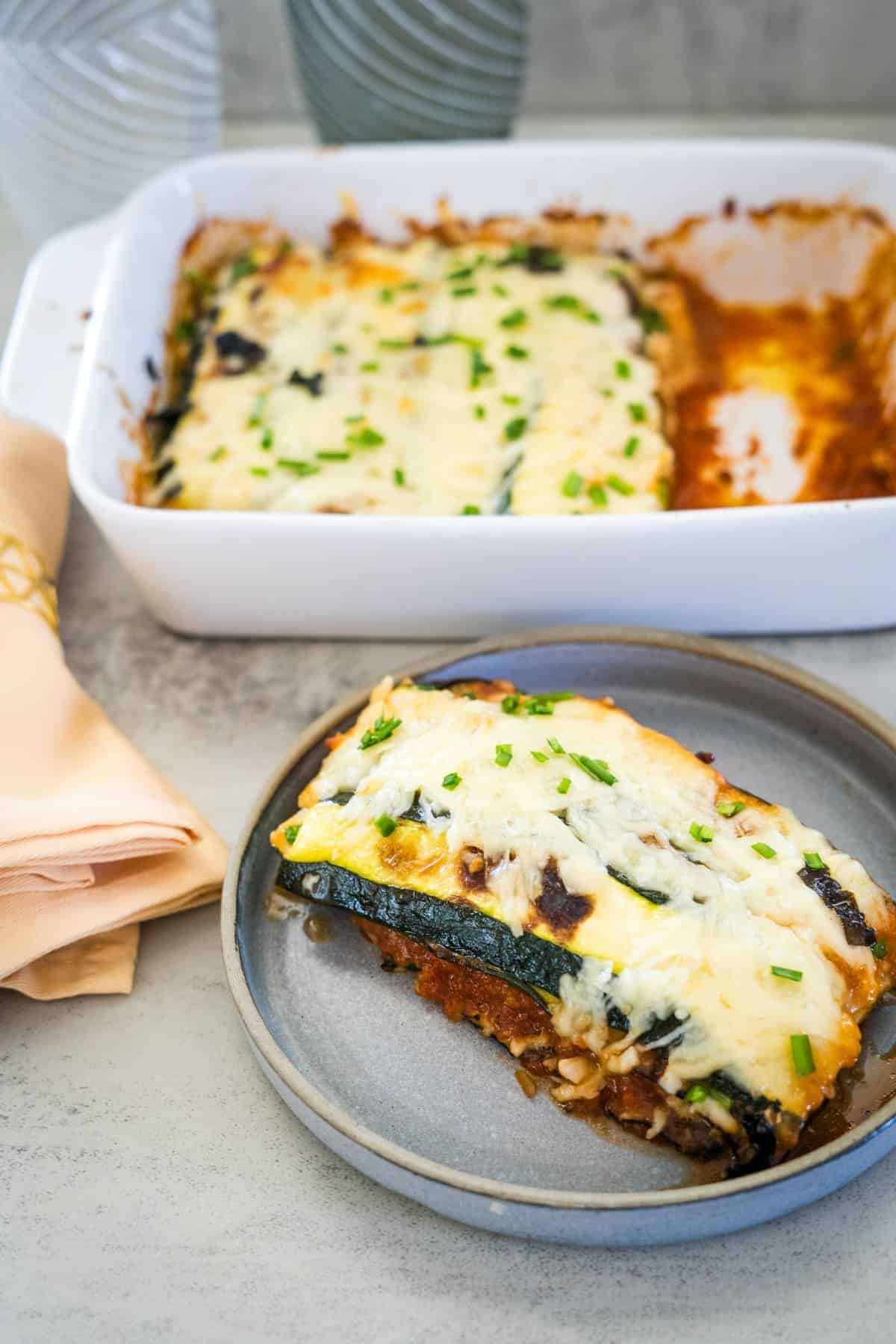 A serving of zucchini parmigiana lasagna rests on a gray plate beside a napkin, with the remaining dish in a white baking tray in the background. The lasagna is topped with melted cheese and chopped herbs.