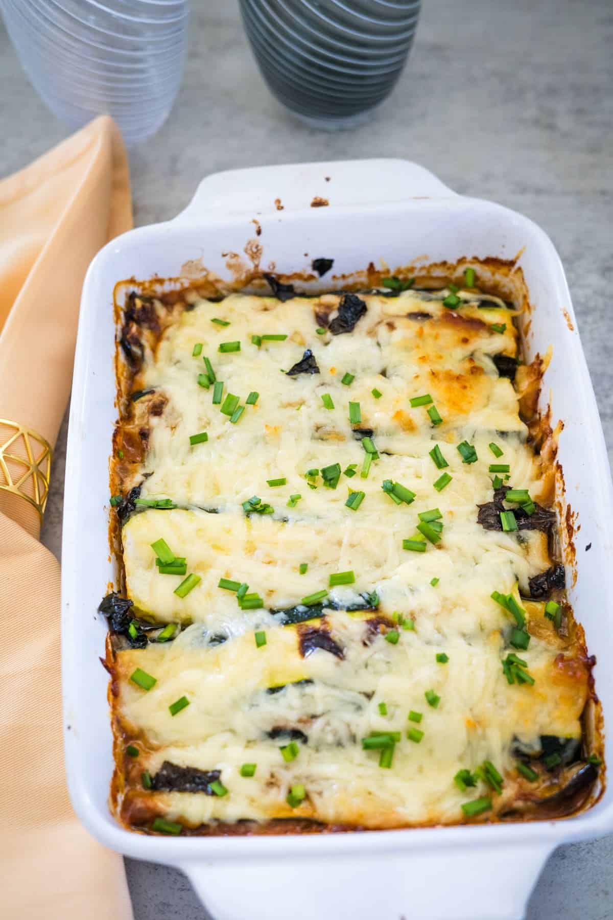 A white rectangular baking dish filled with a cheesy baked casserole, garnished with chopped green herbs, placed on a table with an orange napkin and two grey glass vases nearby.