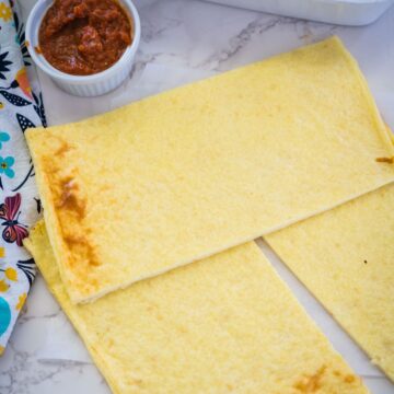 Two rectangular pieces of cooked chickpea flour flatbread next to a small white bowl of red sauce on a marble countertop with a colorful cloth beside them.