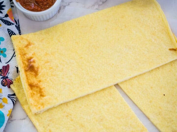 Two rectangular pieces of cooked chickpea flour flatbread next to a small white bowl of red sauce on a marble countertop with a colorful cloth beside them.