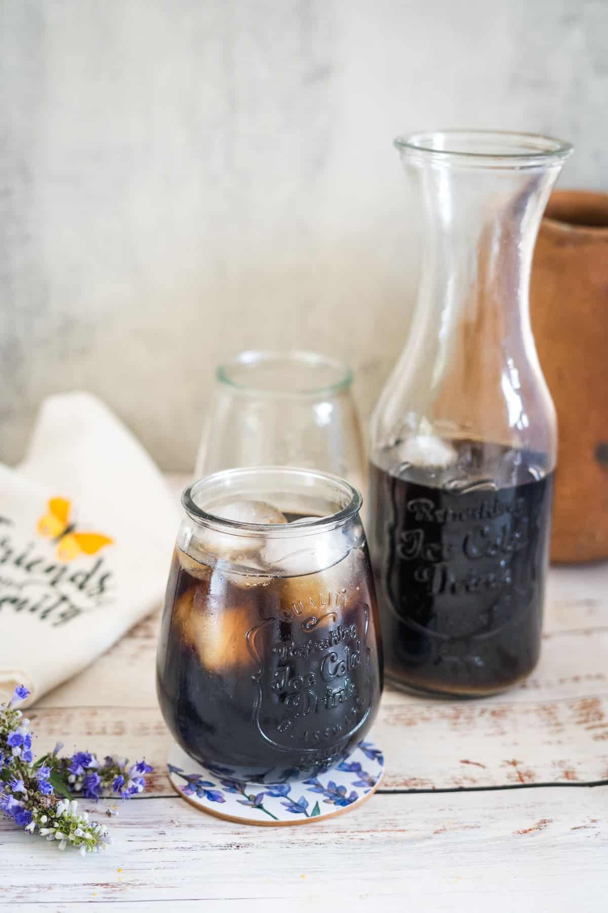 A glass and a carafe filled with iced coffee sit on a wooden surface. An empty glass and a white cloth with text are in the background. Lavender flowers are placed beside the glass.