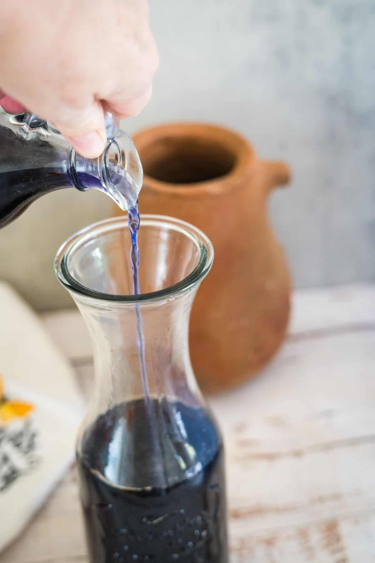 A hand pours a dark blue liquid from a small glass pitcher into a larger glass carafe. An earthenware jug is in the background.