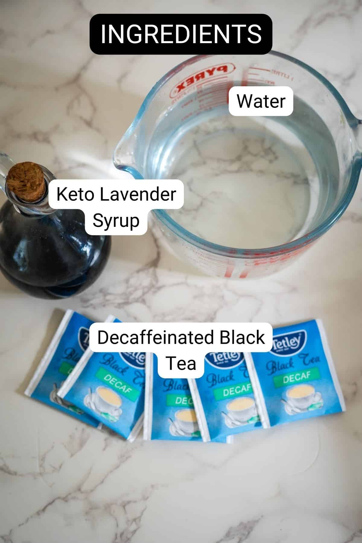 Image showing ingredients for a recipe: a glass pitcher filled with water, a dark bottle labeled "Keto Lavender Syrup," and four sachets of decaffeinated black tea.