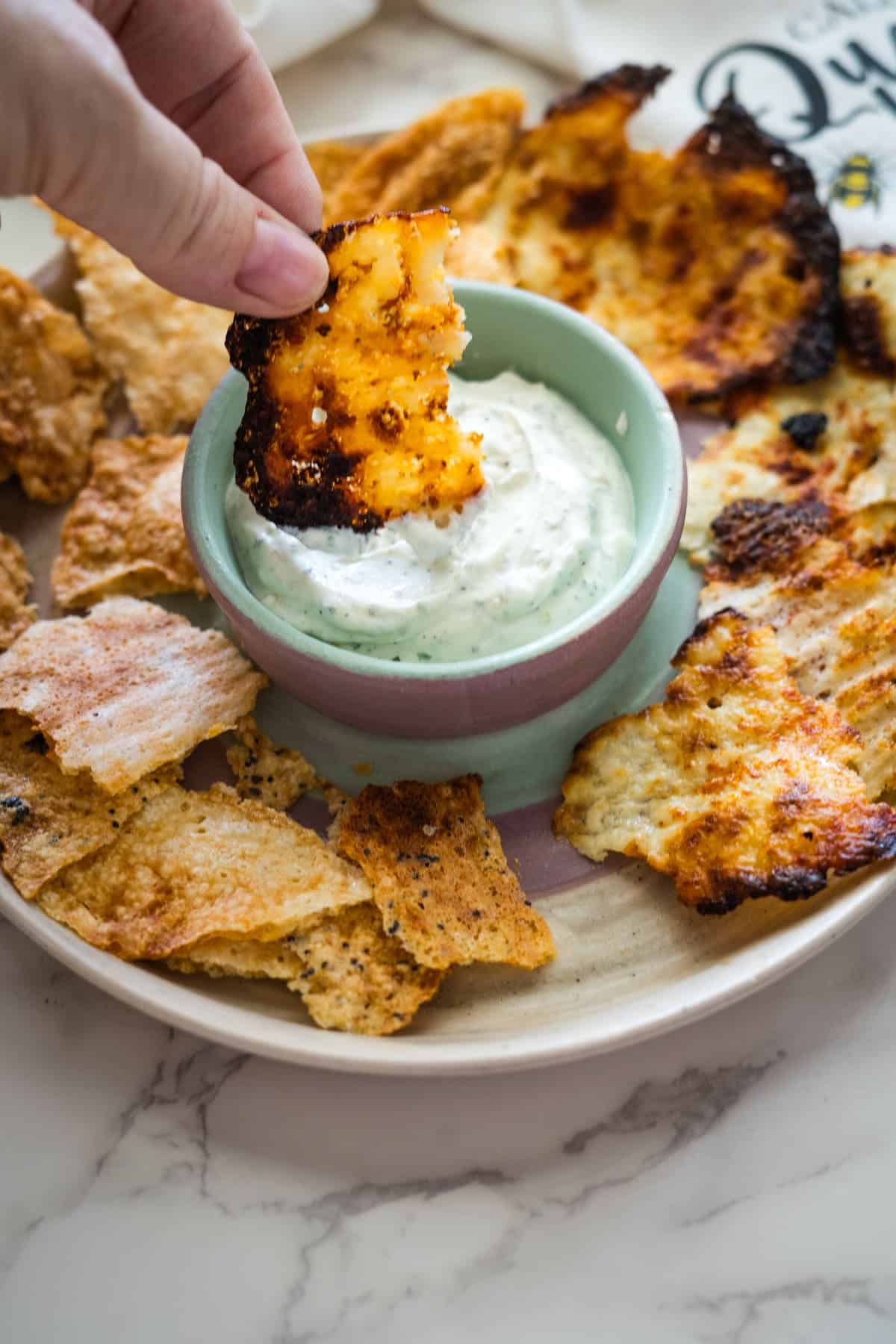 A hand dips a crispy snack into a bowl of creamy dip, surrounded by other similar snacks on a plate.