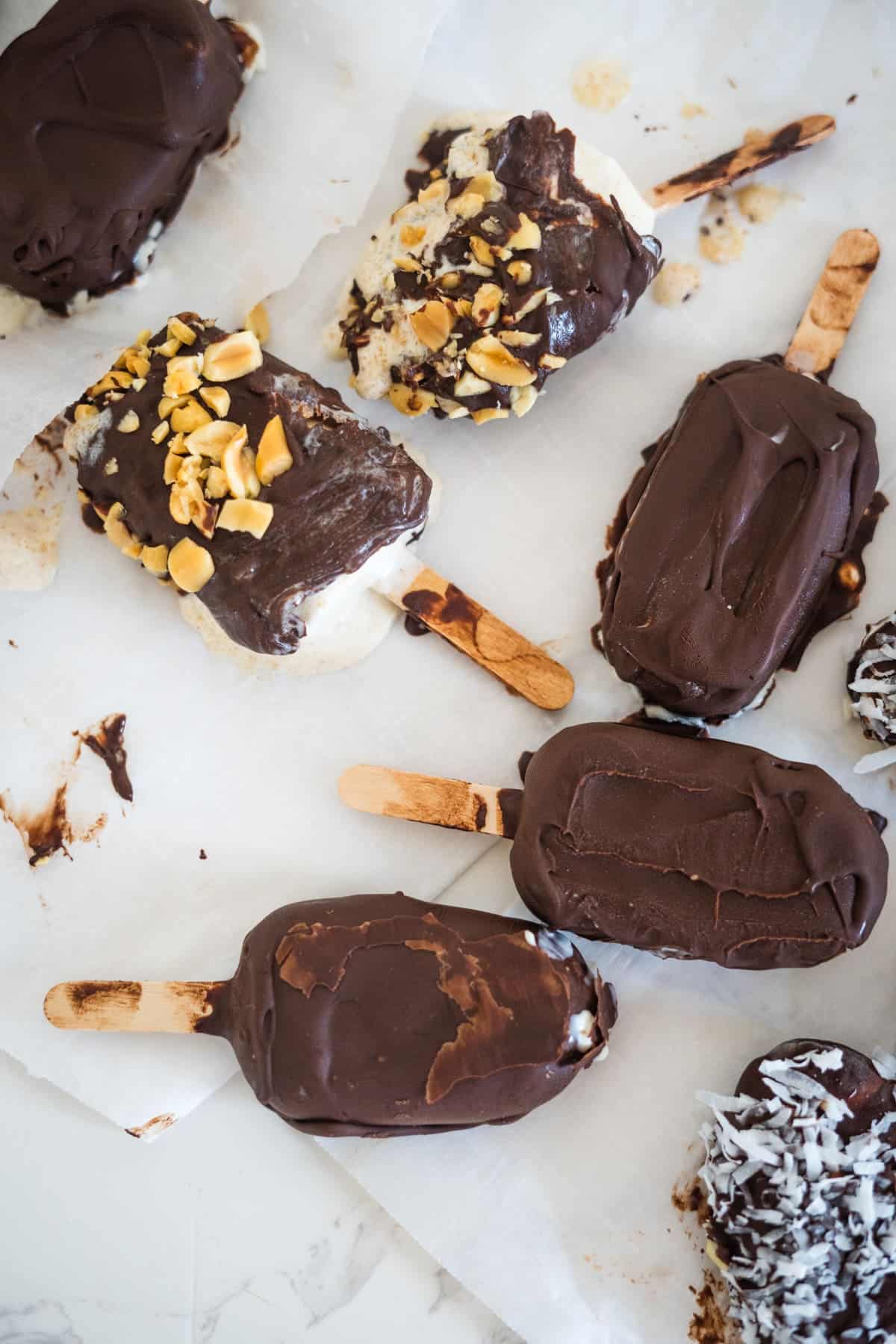 A variety of chocolate-covered ice cream bars, some topped with chopped nuts and shredded coconut, arranged on wax paper.