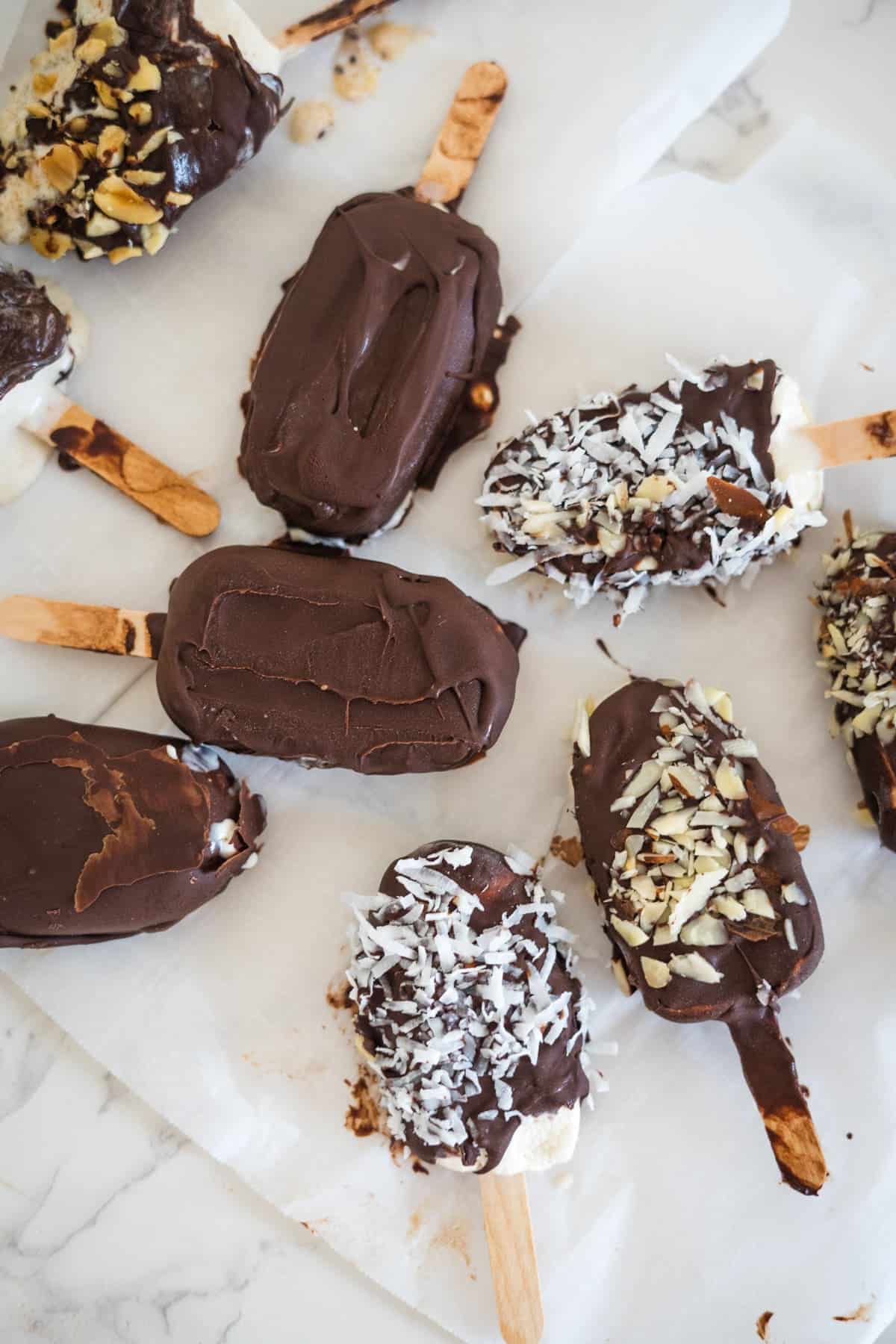 A variety of keto ice cream bars covered in chocolate and topped with nuts and coconut flakes are placed on parchment paper.