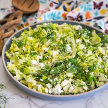 A bowl of chopped salad containing green leafy vegetables and crumbled white cheese, placed on a white marble surface with a colorful patterned cloth and wooden utensils in the background.